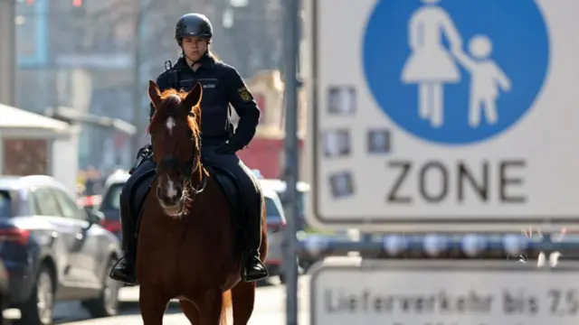 A police officer on a horse patrols the area near the scene of the incident; a zone sign showing a mother and child can be seen in the background..