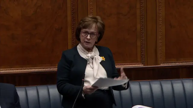 Diane Dodds eading from a piece of paper in the chamber. She has short brown hair and is wearing pink rimmed glasses. She is wearing a dark jacket with a cream top and has a yellow daffodil pin badge on her chest.