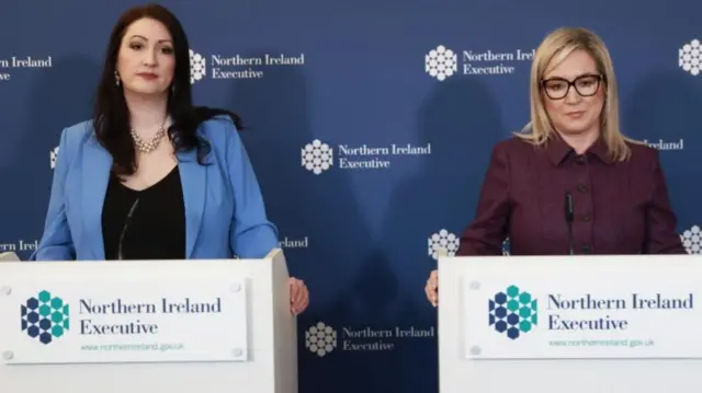 Emma Little-Pengelly and Michelle O'Neill both stand behind separate Northern Ireland Executive branded stands as look towards the camera. The background is also executive branded and has a blue background.
