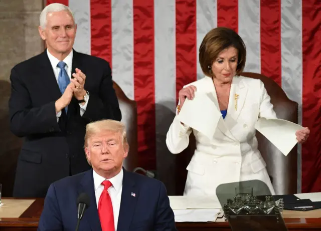 Trump in a red tie speaking at a microphone with Pence behind him clapping and Pelosi in a white suit tearing paper