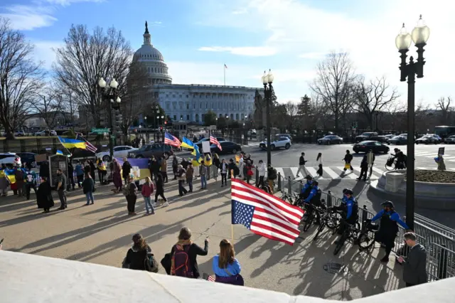 Capitol dome against blue sky with clouds, protesters in foreground with security fencing and police on bikes