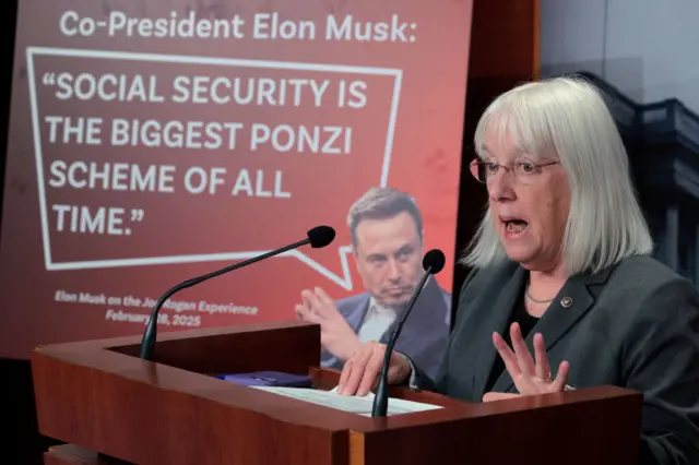 Murray at lectern with microphones in front of poster reading "Co-President Elon Musk: Social Security is the biggest Ponzi scheme of all time"£