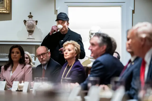 Cabinet members and Donald Trump sit at a table; Elon Musk in black baseball cap stands behind sipping from a cup