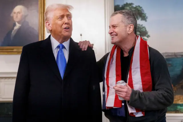 Trump in a blue tie in front of a portrait of George Washington, with Fogel wearing a flag draped around his neck putting his hand on Trump's shoulder