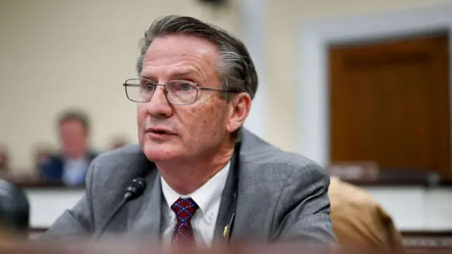 Tim sitting at a desk, which is blurred. He speaks into a small microphone that is on a stand. He wears glasses, a grey suit, red and blue tie and has grey swept back hair