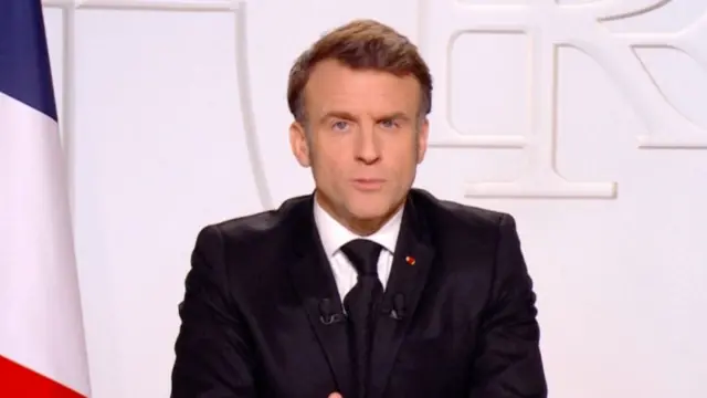 French President Emmanuel Macron giving a televised address in front of a white background next to the French and European flags