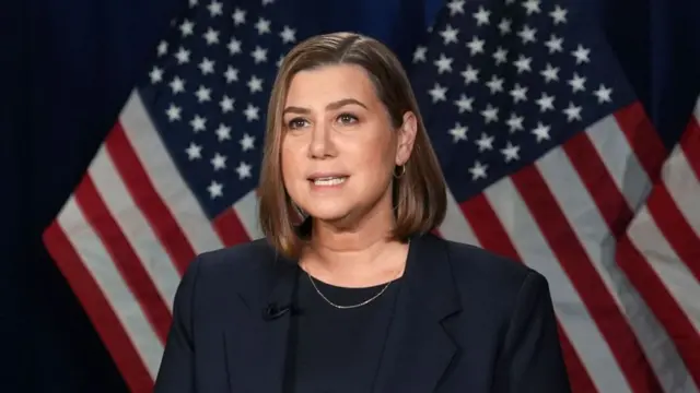 A woman speaking in front of two US flags. She wears a black suit and top and had a short brown bob.