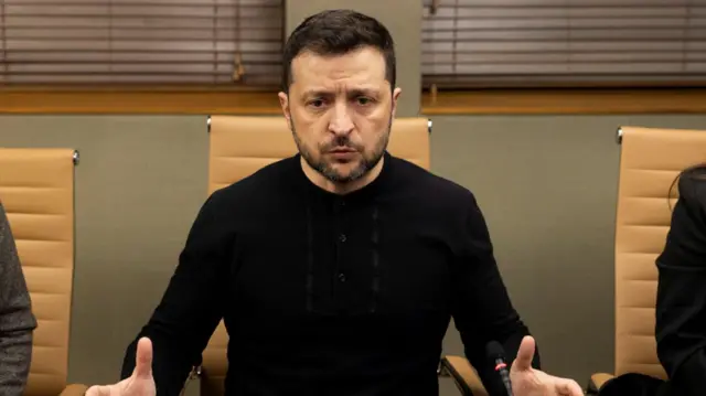 Volodymyr Zelensky sitting down on beige leather swivel chair as he speaks to reporters in a grey-walled room at Stansted Airport. He's wearing a black long-sleeved shirt