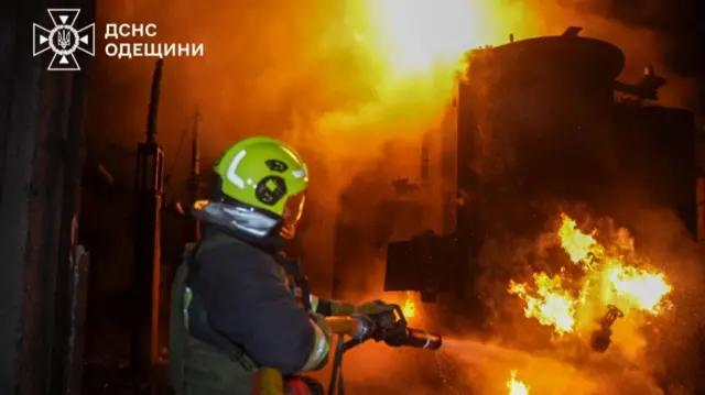 A firefighter in the middle of a fire, using a hose against a big blaze. You can't tell the structure the fire is engulfing but it looks industrial