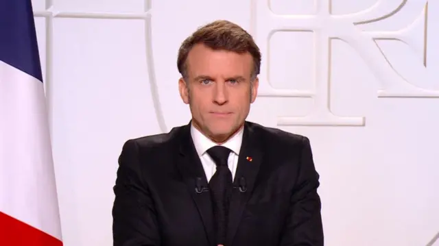 Macron looks to camera during his address. He is wearing a suit and a French flag is visible behind him