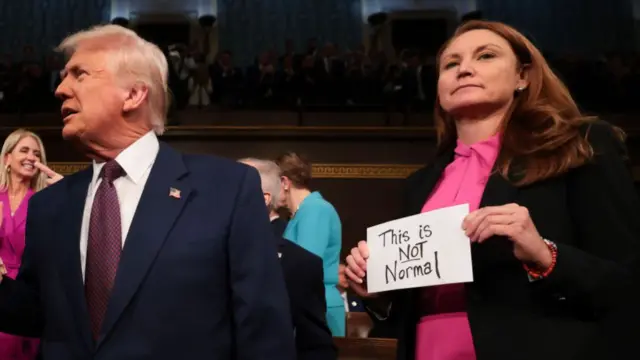 A woman standing on the left of Donald Trump, wearing  a pink  blouse and black blazer. She looks at the camera holding a sign that reads this is not normal. Trump is looking the other way in conversation with someone out of view