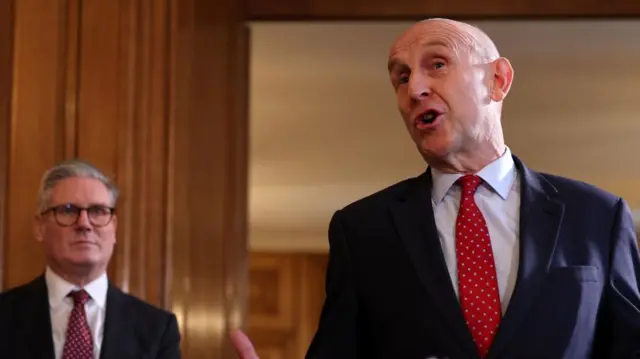 Hesgeth speaking. In the background Starmer watches on as he stands in front of a wooden wall