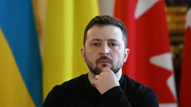 Zelensky looking straight at the camera wearing a black jumper. He holds one hand on his chin. There is a draped Ukrainian flag behind him and a red and white one