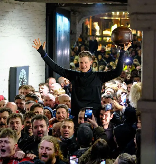 A man on someone's shoulders carrying the Shrovetide ball in a crowd of people