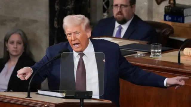 Donald Trump delivers speech in Congress wearing a dark blue suit, maroon tie and white shirt. He's standing behind a lectern, a thick black binder on top of it, a female (L) and a male (R) clerks behind him