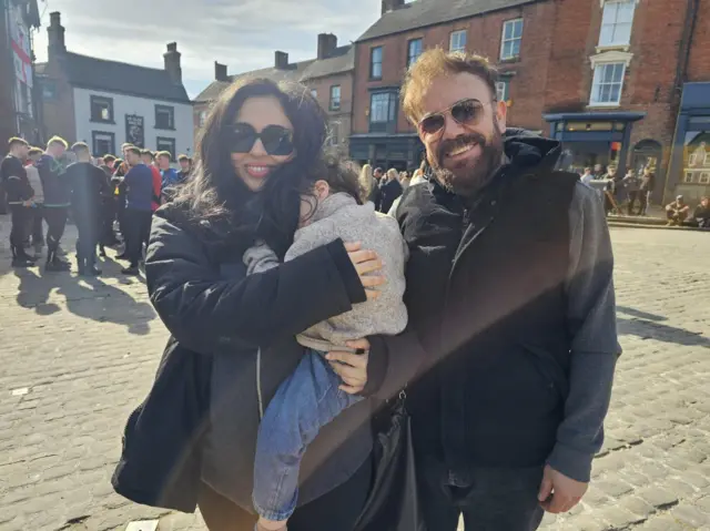 Oumkaltoum Laghrissi holding her son, stood next to her husband. They are both wearing sunglasses and coats.