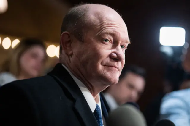 Coons in a dark suit looking to the side with people and lights in the background