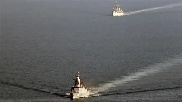 A grey sea. Two large grey ships sail in it, one is ahead and one trails behind