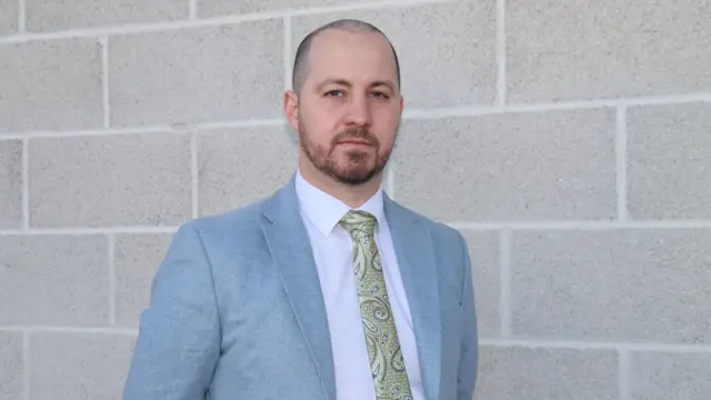 Det Ch Insp Nick Gardner standing in front of a breeze-block wall. He is wearing a light blue jacket and a patterned tie