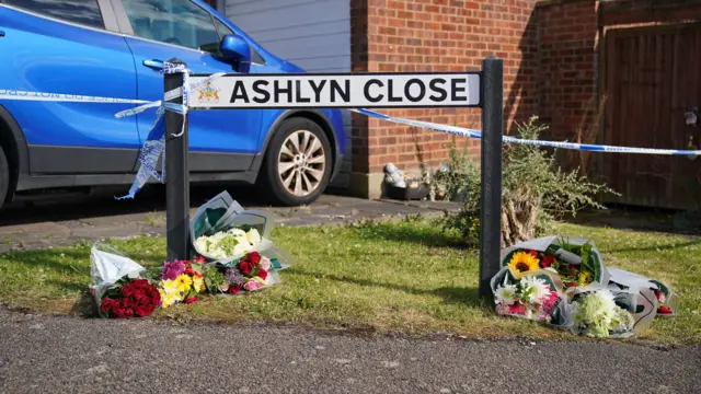 A road sign reading Ashlyn Close. Bouquets of flowers are on the grass. A blue car is parked behind, beside a garage