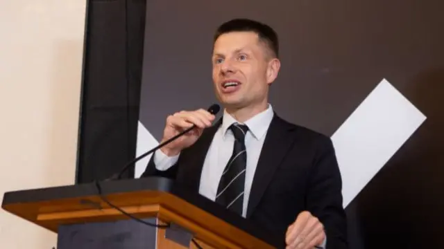 Oleksiy Goncharenko in a suit speaking at a podium into a small microphone which he holds with one hand. He rests the other on the podium