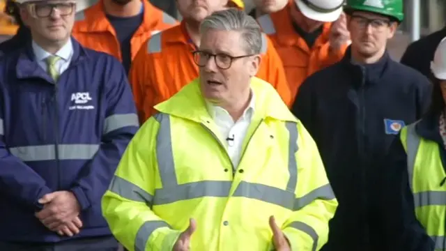 Starmer stood in a high vis jacket as he speaks, with his hands in front of him. People stand behind him in uniforms.