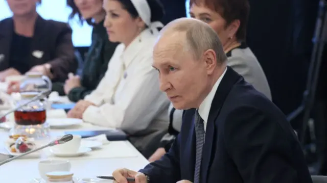 Vladimir Putin at a meeting wearing a suit, white shirt and tie.