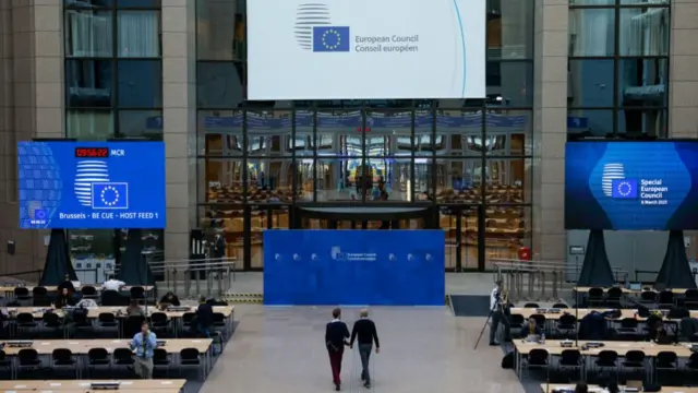 A huge room with a tall ceiling and large screens with dozens of chairs in a row and two men walking through