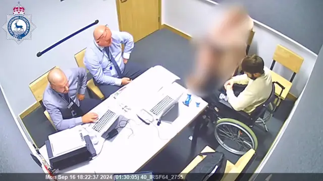 A CCTV ariel shot of an interrogation room with two police officers by a desk with laptops, and Kyle Clifford in a wheelchair head down. A fourth person is blurred out.