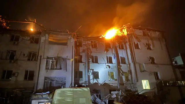 Fire can be seen around the roof of a building, which has been damaged. The picture has been taken at night-time.