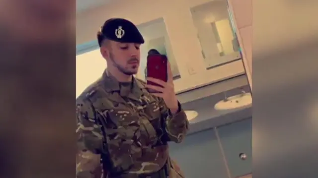 Selfie of Kyle Clifford wearing his army uniform and beret in a bathroom. He is holding a red phone and has a dark, close-cropped beard and moustache.