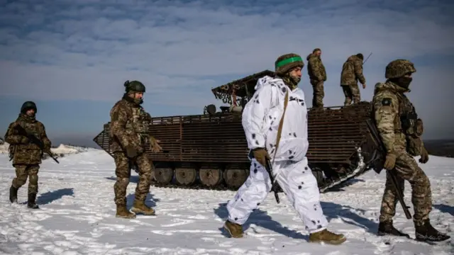 Ukrainian soldiers practice firing from a gun of the BMP-1 infantry fighting vehicle. The group of six men - five in regular green military uniform, one at the front in white winter uniform - walk alongside the vehicle in the snow