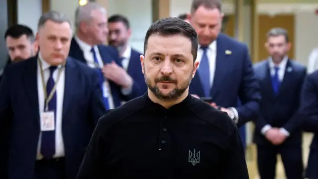 Ukrainian President Volodymyr Zelensky wears a black top with various officials in the background.