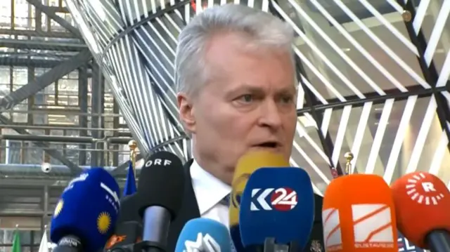 A grey haired man speaking into several press microphones pressed around him covering some of his face