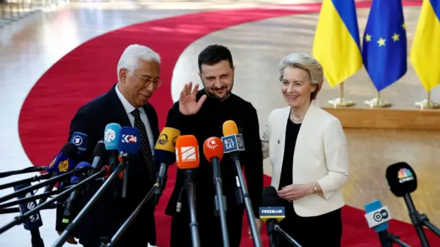 Costa (L), Zelensky (C) and von der Leyen (R) stand in front of mics as they address the press. Zelensky waves with his right hand