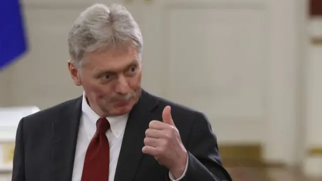 Dmitry Peskov stands in black suit, red tie and white shirt as he holds up his left hand showing a thumbs up.