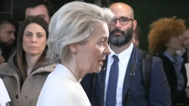 From the side, Leyen speaks to a press microphone. She wears a white jacket and has a short blonde bob