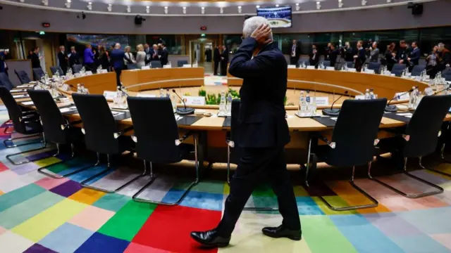 A large round table with empty chairs, European Council President Antonio Costa is walking past the table while press and other leaders huddle in the distance