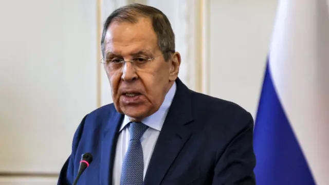 Close up of Sergei Lavrov standing at lectern in dark blue suit, a Russian flag partially visible to his left