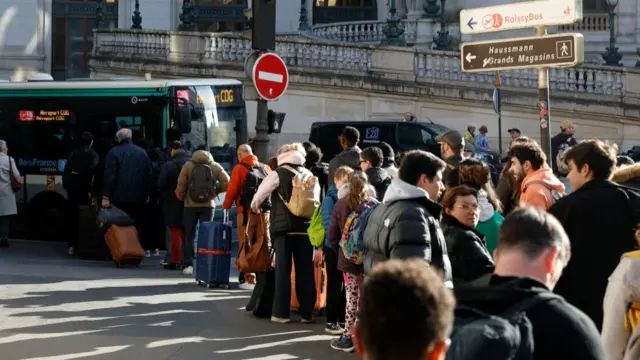 People stand outside with their luggage in a queue for a bus