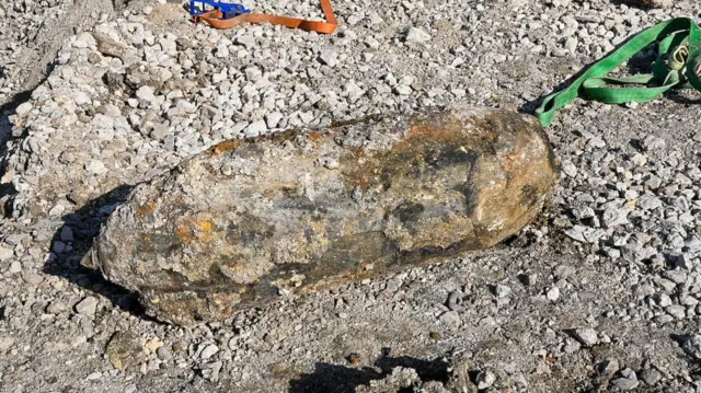 Unexploded WW2 bomb covered in layers of mud and rubble surrounded by gravel and dirt near rail tracks.