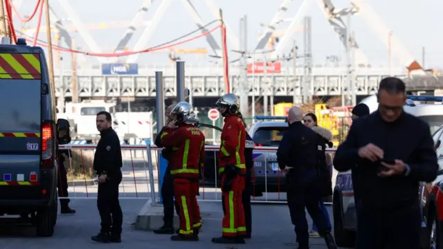 Emergency service workers, some in high vis, stand in front of a barrier, with the freight area in Saint-Denis in the background