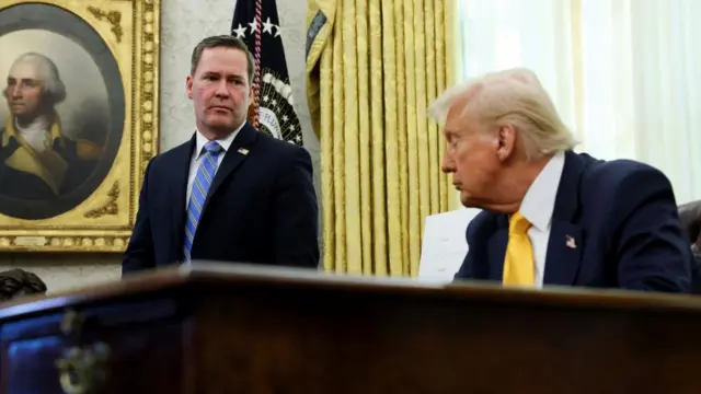 Mike Waltz in a blue tie, standing in the Oval Office. Donald Trump sat in front of him, behind a large wooden desk.