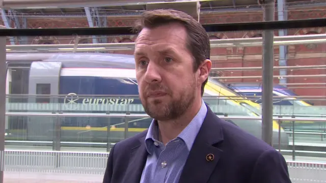 Simon Lejeune speaking to camera. A Eurostar train is visible in the background