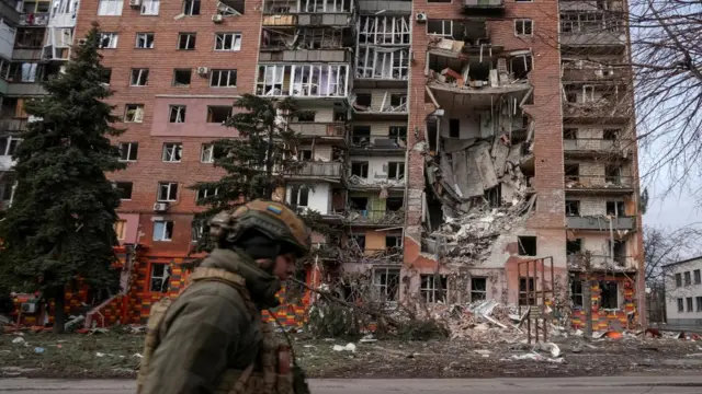 Soldier walks in front of a large high-rise building, with rubble on the ground and a large hole in the building with damage to the inside of the building.