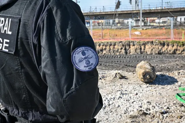 An officer in jacket stands facing away from camera. Unexploded bomb can be seen in front of him with tracks and fence visible.