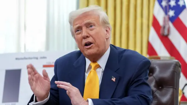 Trump gestures as he speaks sitting at a desk, with the US flag behind him