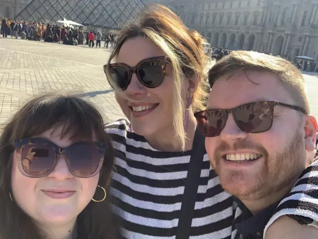 Two women and a man smile for a selfie outside the Louvre museum. All three wear sunglasses.