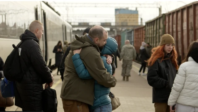 Two people embrace one another in a hug, as they stand on a train platform with many people standing around them. One of the people, a man, is in military fatigue, while the person he's hugging is in plain clothes.