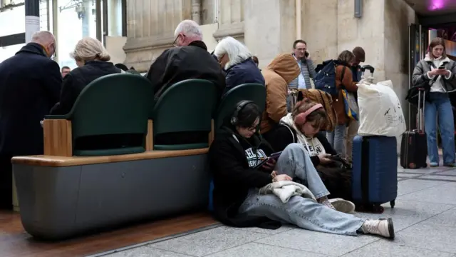 Two female passengers sit on the floor, with headphones on, behind a group of seats filled with travellers.